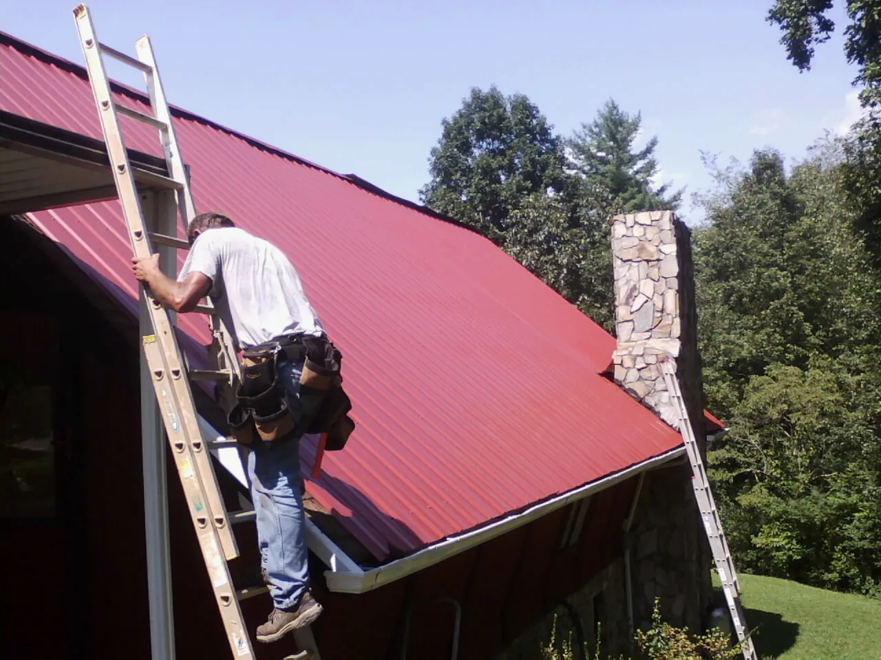 Roofer on a ladder beside a red metal roof and stone chimney — Soffit & Fascia Repair by Roofing Brothers in Idyllwild